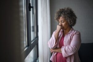 An older woman standing by a closed window with her fist on her mouth and her eyes downcast, showing she's sad or depressed